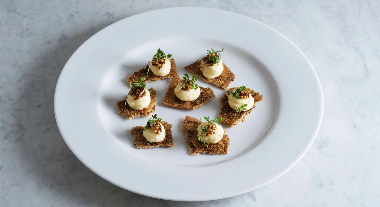 Ett smakfullt portionsupplägg med krämig ädelostspread på julkryddat knäckebröd. De syltade senapsfröna med lite sötma och knaprig textur är ett enkelt, imponerande garnityr.