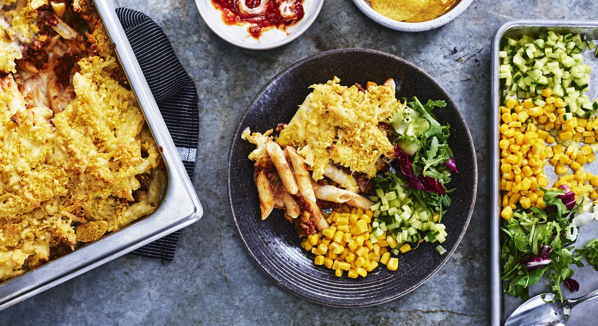 Den här pastagratängen har tacokryddad köttfärs och majs under gratängtäcket av ost och krossade nachos. Tacosmaken är populärt bland eleverna på skolan så det här kommer att bli en favoriträtt i lunchmatsalen. Servera gärna med yoghurt blandad med het salsa, majs, gurka och grönsallad.