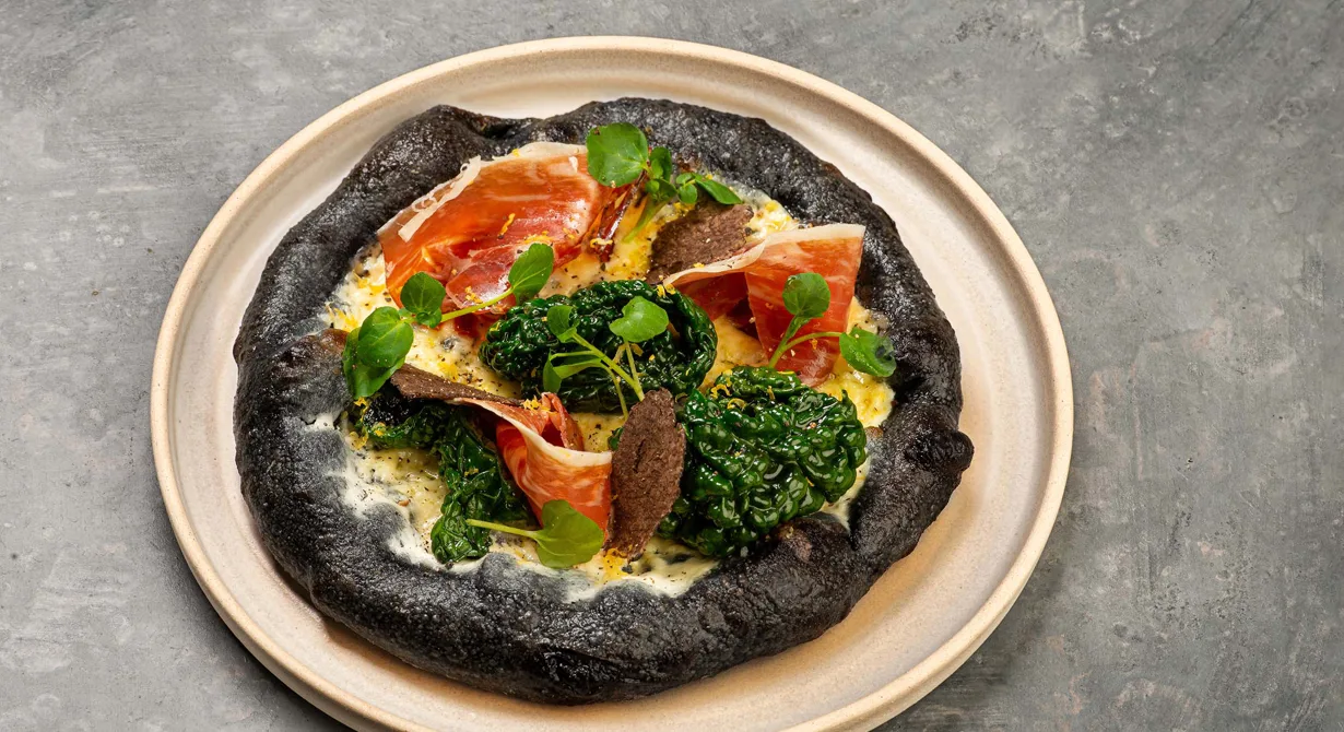Den svarta pizzan, färgad med aktivt kolpulver, är faktiskt en pizza bianco då den täcks med crème fraiche och långlagrad Präst®. Svartkålen går med i pizzaugnen och får en krispig textur. Pizzan toppas därefter med pata negra, tunt hyvlad svart tryffel och vattenkrasse.